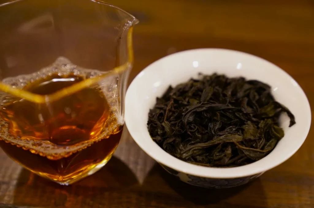 A glass pitcher with amber-colored tea and a white bowl filled with dark, dried tea leaves sit on a wooden table, exuding a warm, inviting ambiance.