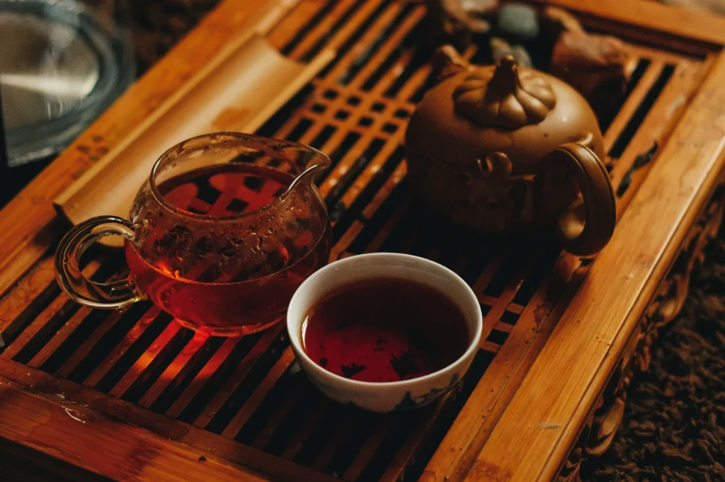 A cozy tea scene with a glass teapot, a ceramic teapot, and a filled teacup on a wooden tray. The warm lighting creates a relaxed ambiance.
