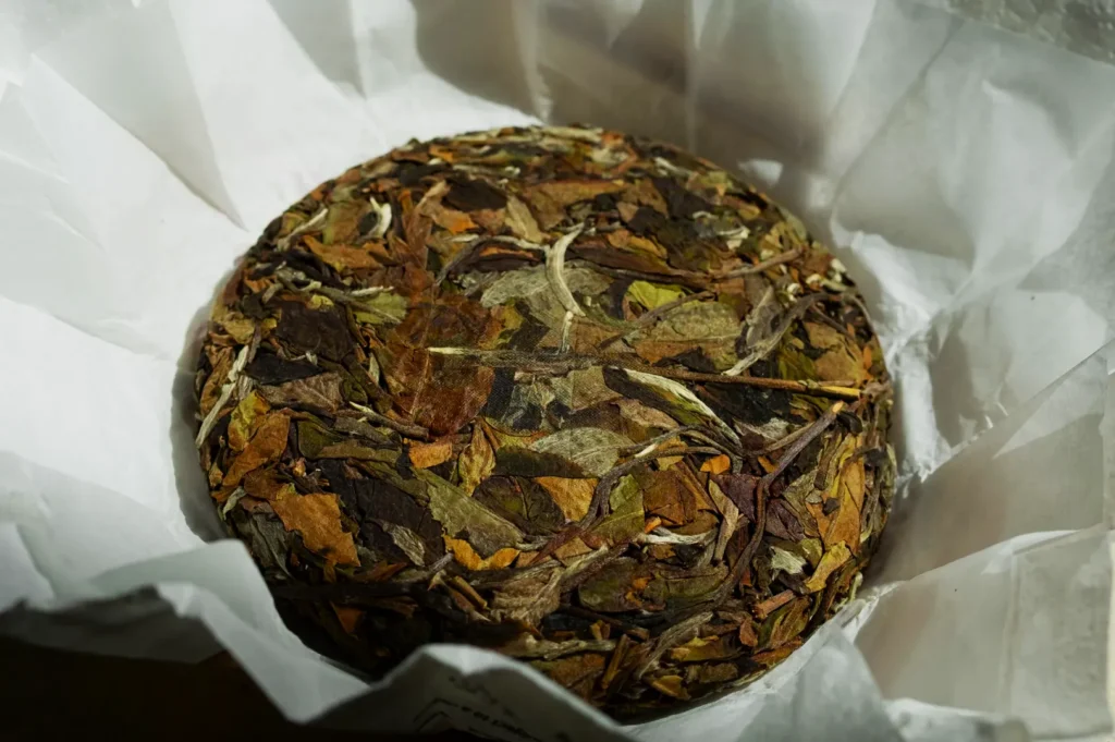 A round, compressed disc of dried tea leaves wrapped in white paper. The earthy tones of the leaves give a natural, rustic impression.