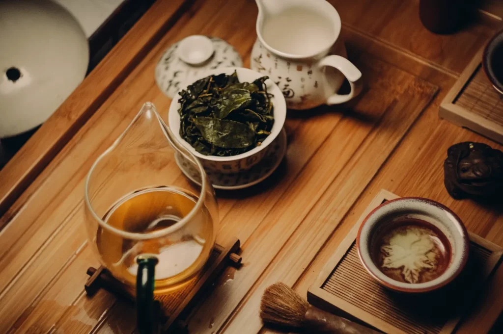 Tea ceremony setup with a glass pot of brewed tea, a bowl of tea leaves, a patterned pitcher, and a small cup on a wooden tray, creating a calming ambiance.
