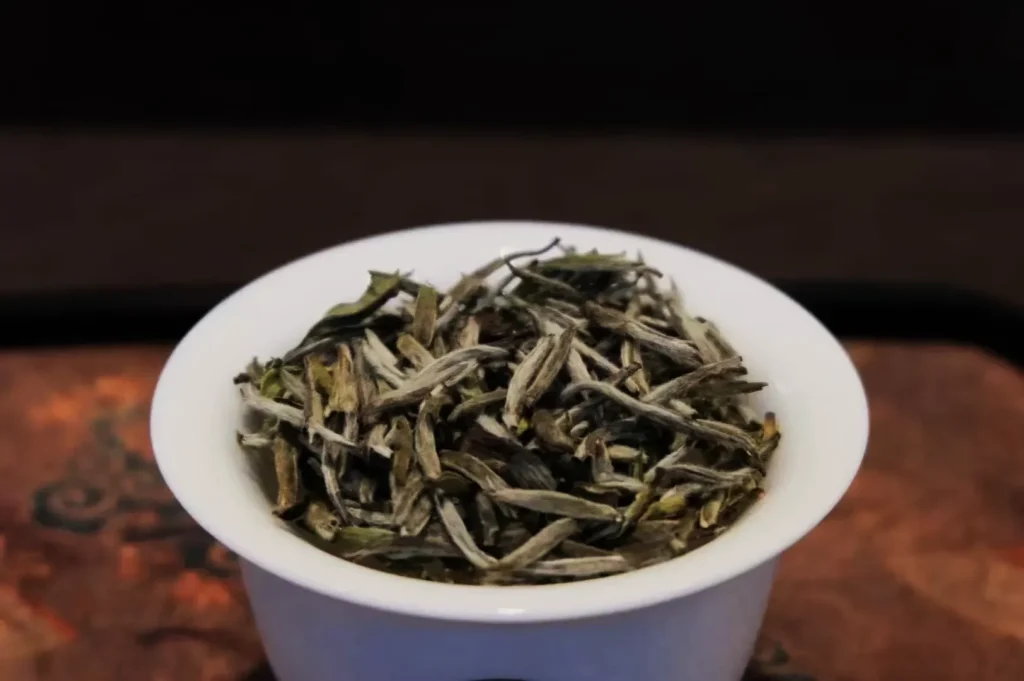 A white bowl filled with dry, slender leaves of white tea sitting on a textured surface, conveying a rustic and natural feel.