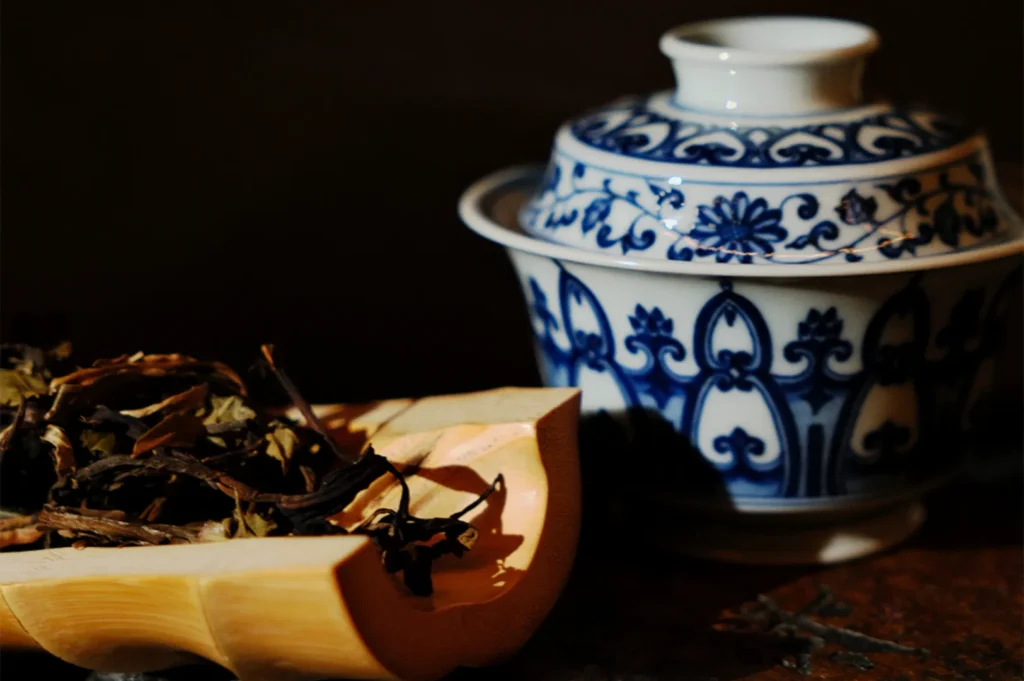 A decorative blue and white porcelain tea cup with lid sits next to a wooden tray filled with dried tea leaves. The scene evokes tranquility and tradition.