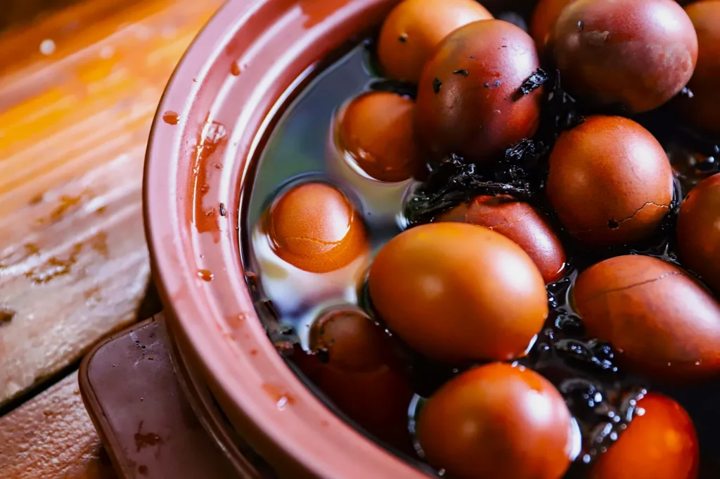 Brown eggs are submerged in dark tea-infused water inside a clay pot, on a wooden surface. The scene gives a rustic, traditional cooking vibe.
