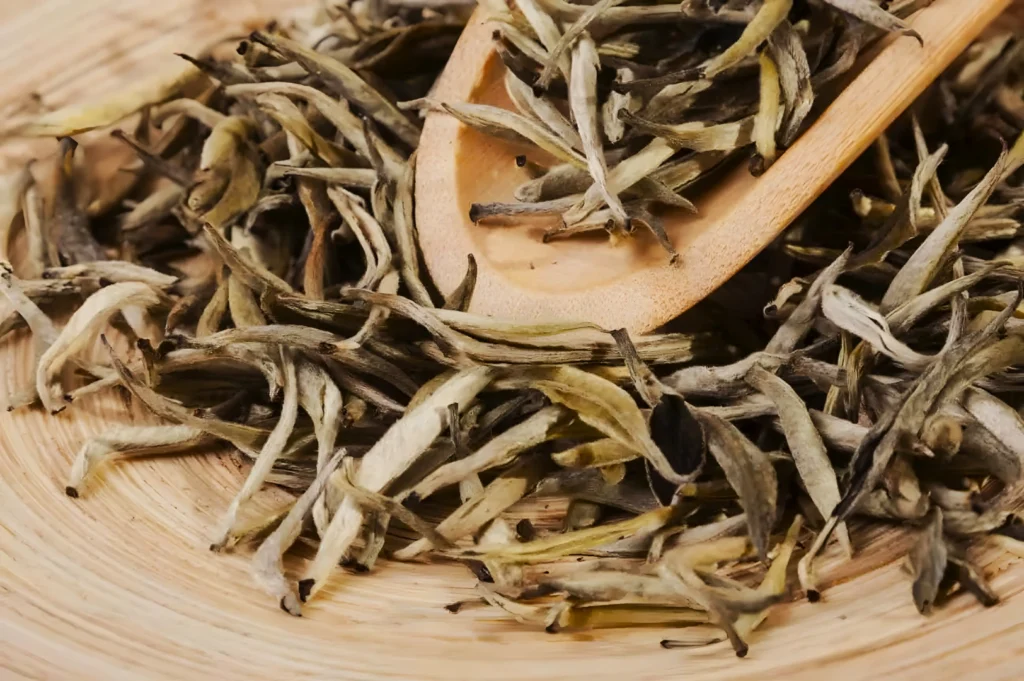 Close-up of dried tea leaves spilling from a wooden spoon onto a woven surface. The leaves are slender and slightly curled, in earthy tones.