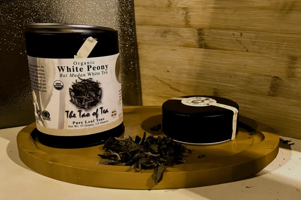 A canister of organic White Peony tea sits on a round wooden tray, accompanied by a smaller tin. Loose tea leaves are scattered nearby, creating a cozy, inviting ambiance.