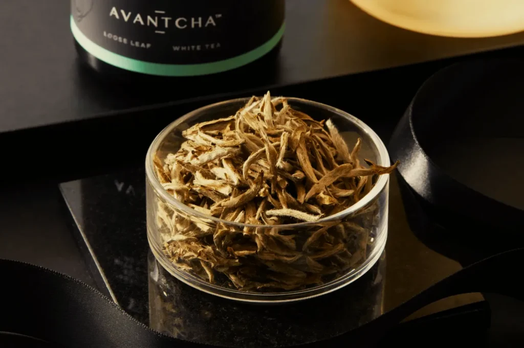 A small glass dish holds loose leaf white tea on a black surface. Behind it, a cylindrical container labeled "Avantcha" sits partially visible, creating a calm, elegant ambiance.
