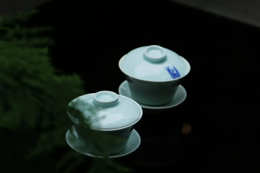 Two white porcelain teacups with lids rest on saucers against a dark background. Soft greenery in the foreground adds a serene touch.