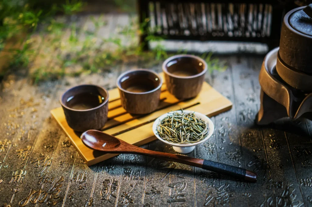 Three brown tea cups on a wooden tray beside a white bowl with dried tea leaves and a wooden spoon. Background features elegant calligraphy.