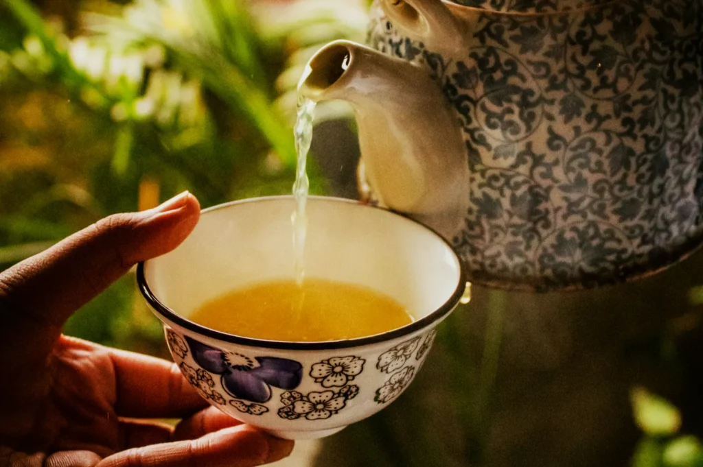 A detailed teapot pours steaming green tea into a floral-patterned cup. The scene is set against a blurred, lush green background, evoking calm and warmth.