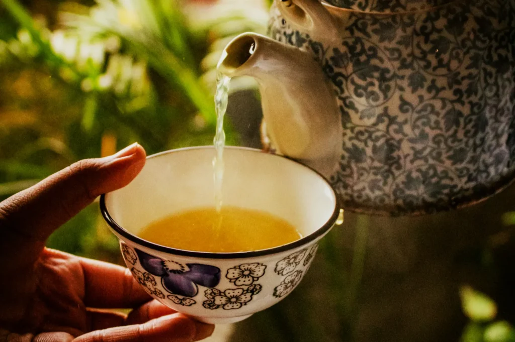 A hand holds a floral-patterned cup being filled with tea from an ornate teapot. The background is blurred greenery, creating a serene atmosphere.