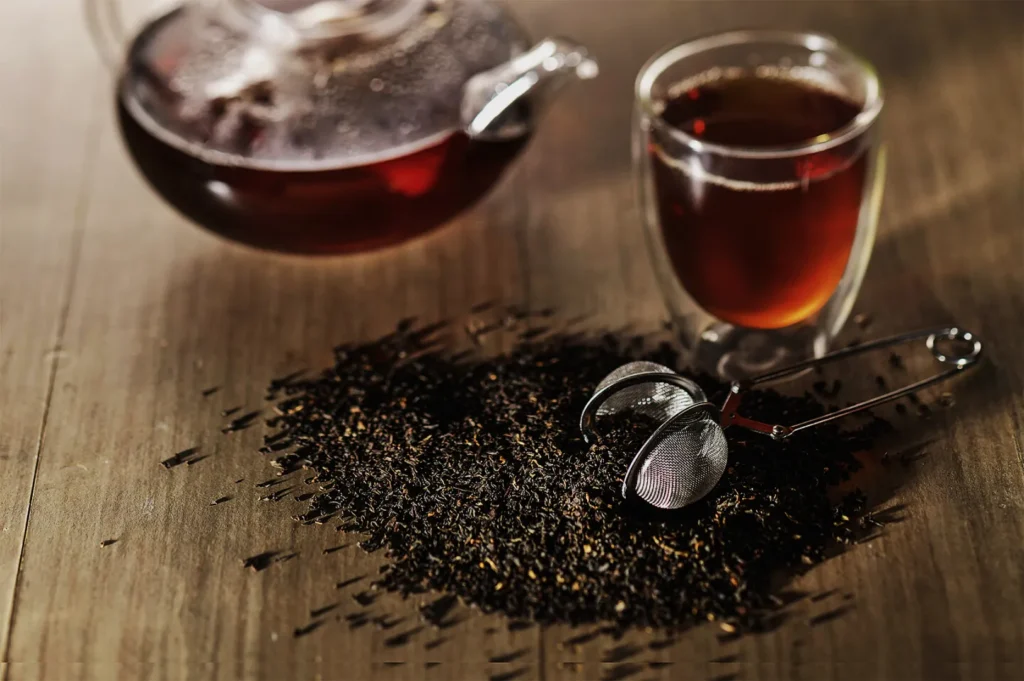 A glass teapot and cup filled with dark tea sit beside loose tea leaves and a mesh tea infuser on a wooden table, evoking a cozy and warm atmosphere.