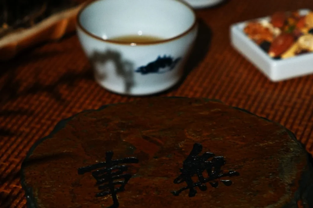 A rustic tea setup features a round mat with Chinese characters, a white teacup filled with tea, and a small dish of mixed nuts, creating a cozy, traditional ambiance.