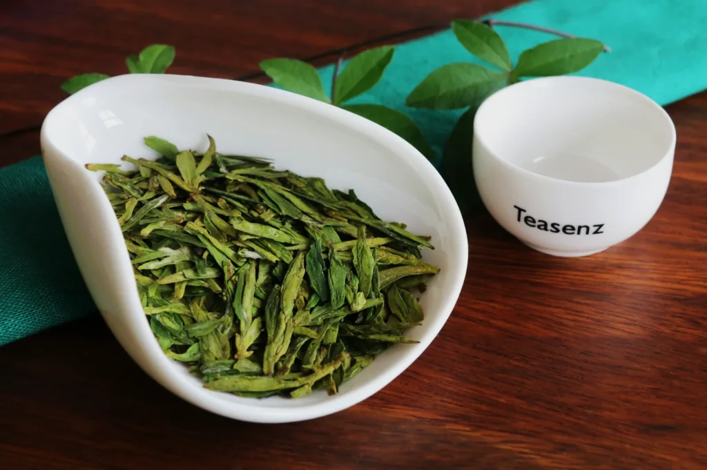 A white dish filled with dried green tea leaves is placed on a wooden table beside a small, empty white cup labeled "Teasenz." A green cloth and leaves add freshness.
