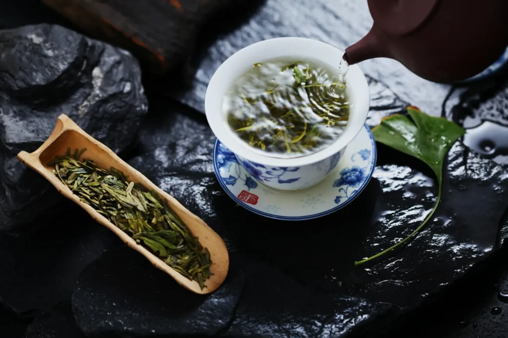 A ceramic teacup with floral patterns is filled with hot water, steeping loose green tea leaves. A wooden scoop holds more dry leaves beside it, creating a serene tea ritual scene. A ginkgo leaf rests nearby on dark slate.