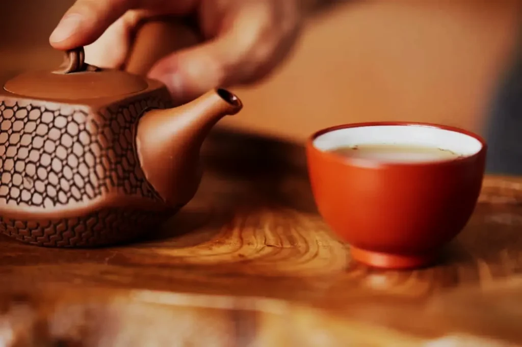 A hand pouring tea from a textured, brown teapot into a matching red cup on a wooden surface, creating a warm and peaceful atmosphere.