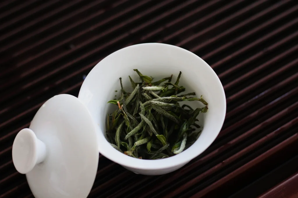 White ceramic bowl with dried green tea leaves, lid ajar, on a dark bamboo mat. The scene evokes calm and anticipation for tea brewing.