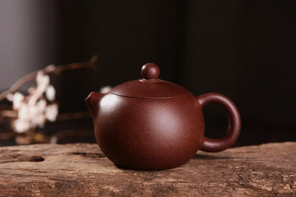A brown clay teapot with a rounded shape sits on a rustic wooden surface. Blurred white blossoms and soft lighting create a serene, traditional ambiance.