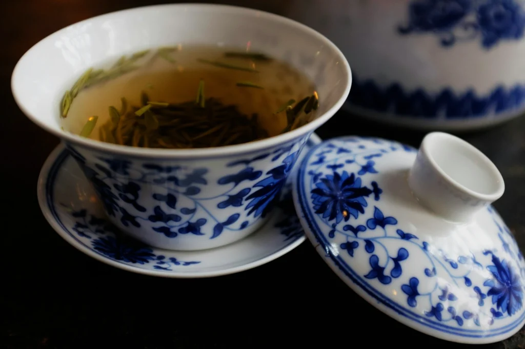 A white porcelain cup with blue floral patterns holds green tea with floating leaves. The matching lid is set slightly ajar, creating an elegant, serene mood.