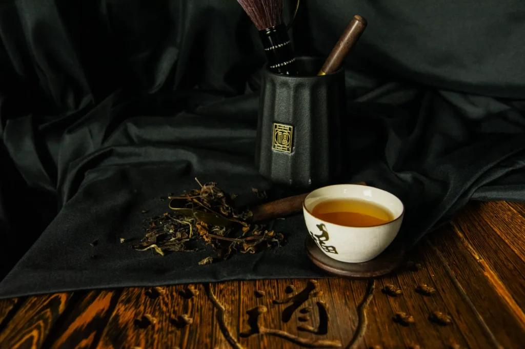 A traditional tea setup on a dark fabric features a white teacup filled with golden tea, dried leaves, and a black pot holding a wooden tea whisk.