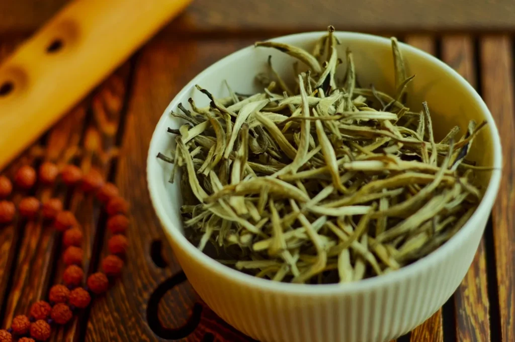 A white bowl filled with long, thin, dried tea leaves sits on a wooden table. Nearby, a string of brown prayer beads adds a serene, meditative ambiance.