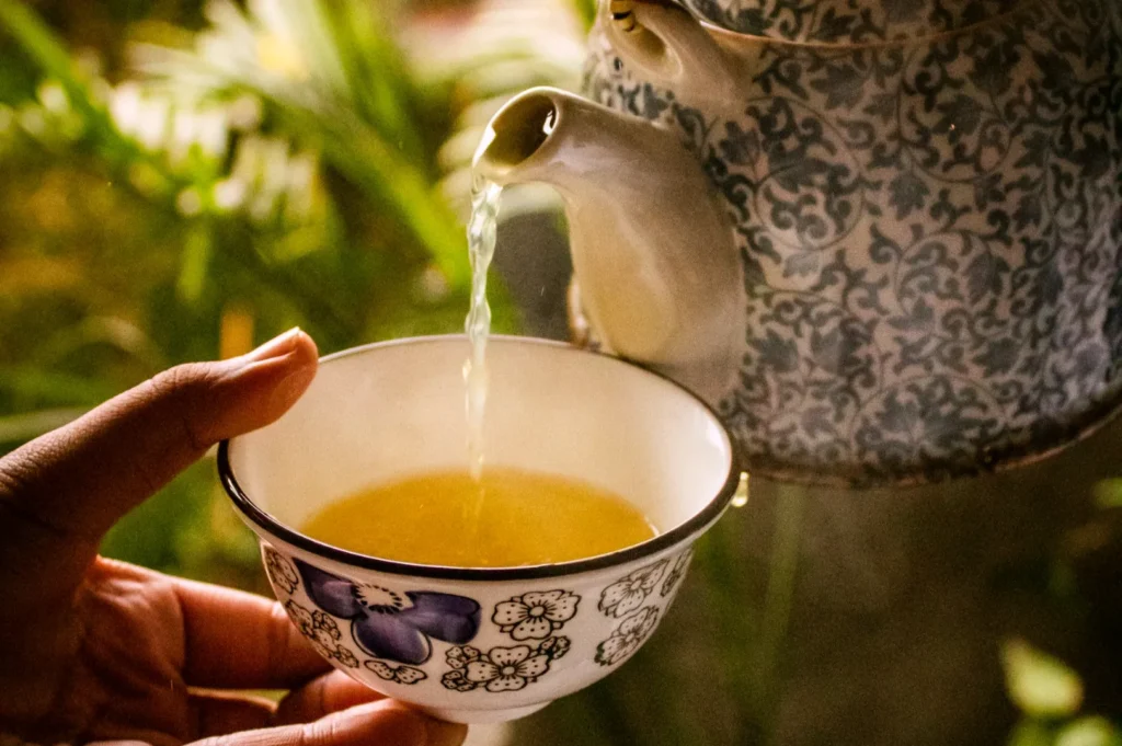 A porcelain teapot with blue floral patterns pours tea into a matching cup with purple flowers. A hand holds the cup amid a warm, cozy setting.
