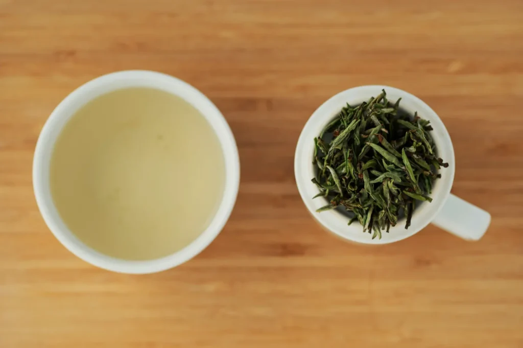Aerial view of a wooden surface with two cups: one holds light green tea and the other contains green tea leaves, conveying a fresh and calming tone.