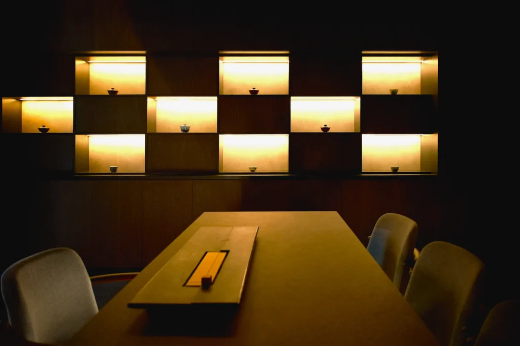 Dimly lit shelves with small pottery pieces are displayed against a dark wall. A minimalist table and chairs create a calm, contemplative atmosphere.