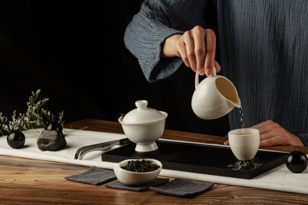 A person in a blue robe pours tea from a white kettle into a small cup on a wooden table. A serene, minimalist tea setup includes a pot, tray, and tea leaves.