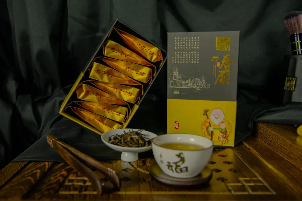 A neatly arranged tea set includes a decorative box of golden sachets, a small white bowl of loose tea leaves, and a cup of tea on a wooden table, evoking a traditional and calming atmosphere.