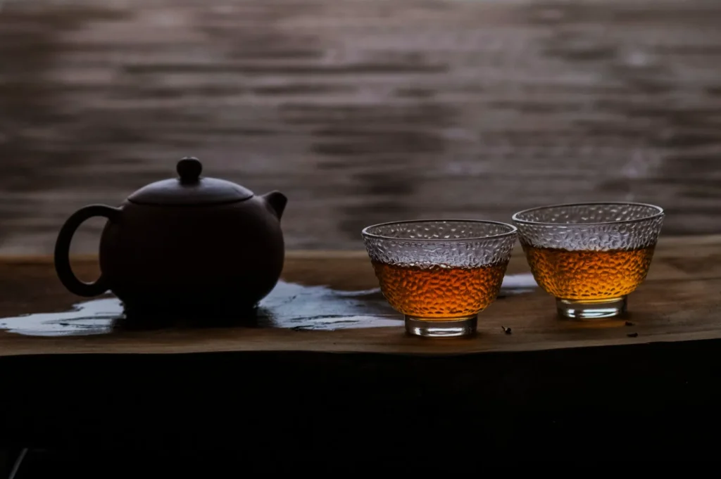 A serene setting with a dark brown teapot and two glass cups of amber tea on a wooden table. The dim lighting creates a calm, reflective atmosphere.