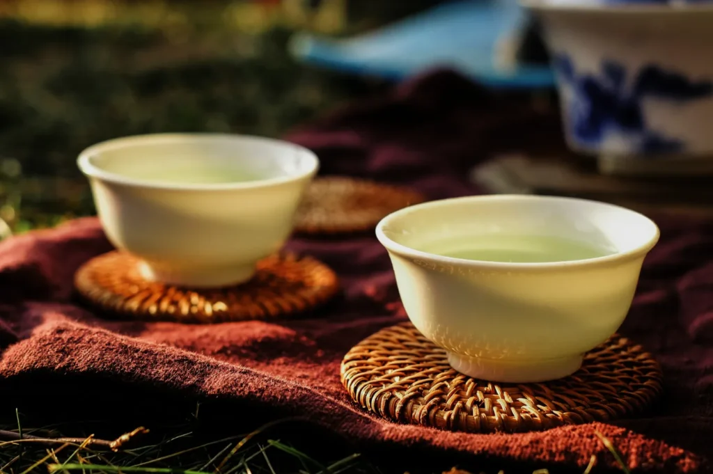 Two white ceramic teacups filled with green tea sit on woven coasters atop a burgundy fabric. The scene is sunlit, evoking a warm, serene ambiance.