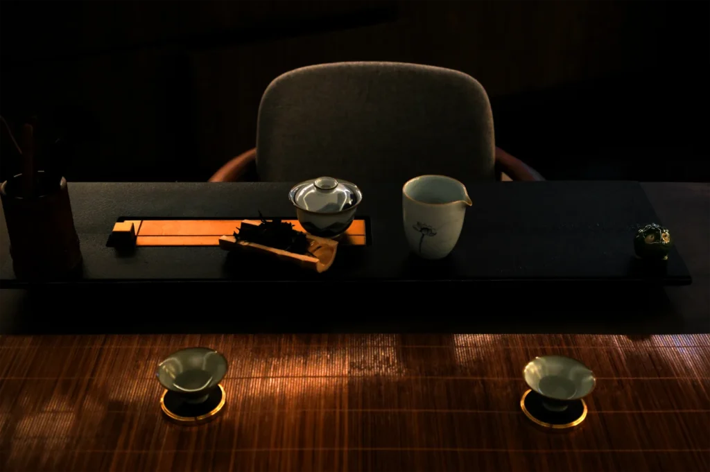Dimly lit tea ceremony setup with a gray chair, teacups, and a teapot on a dark tray. The warm ambiance evokes calm and tradition.