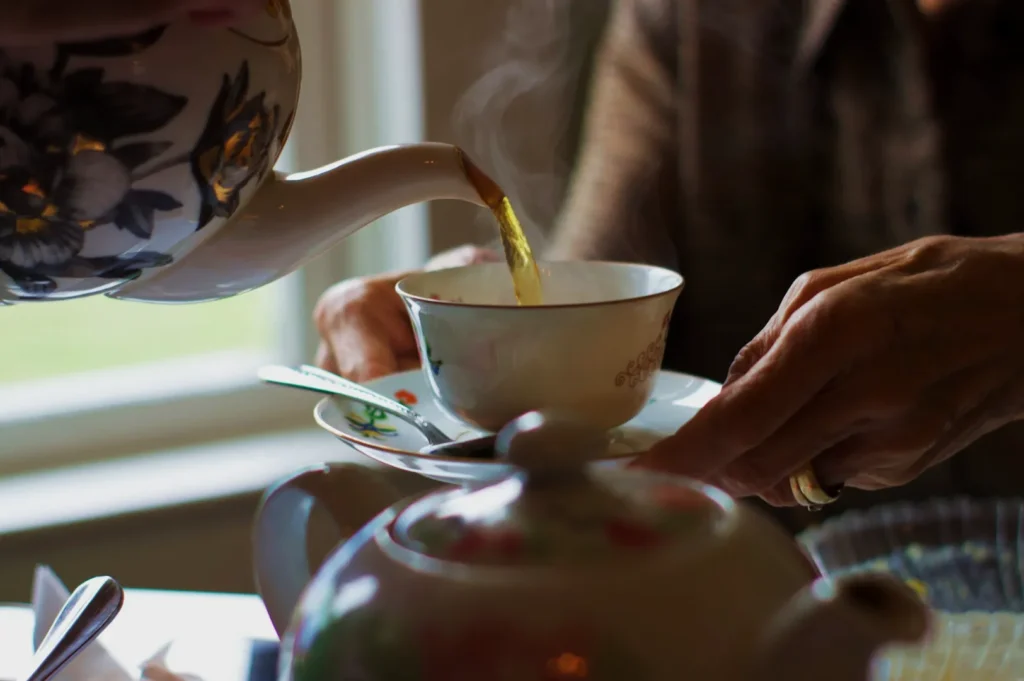 A floral teapot pours steaming tea into a delicate china cup held by an elderly person, creating a warm and cozy atmosphere by a window.
