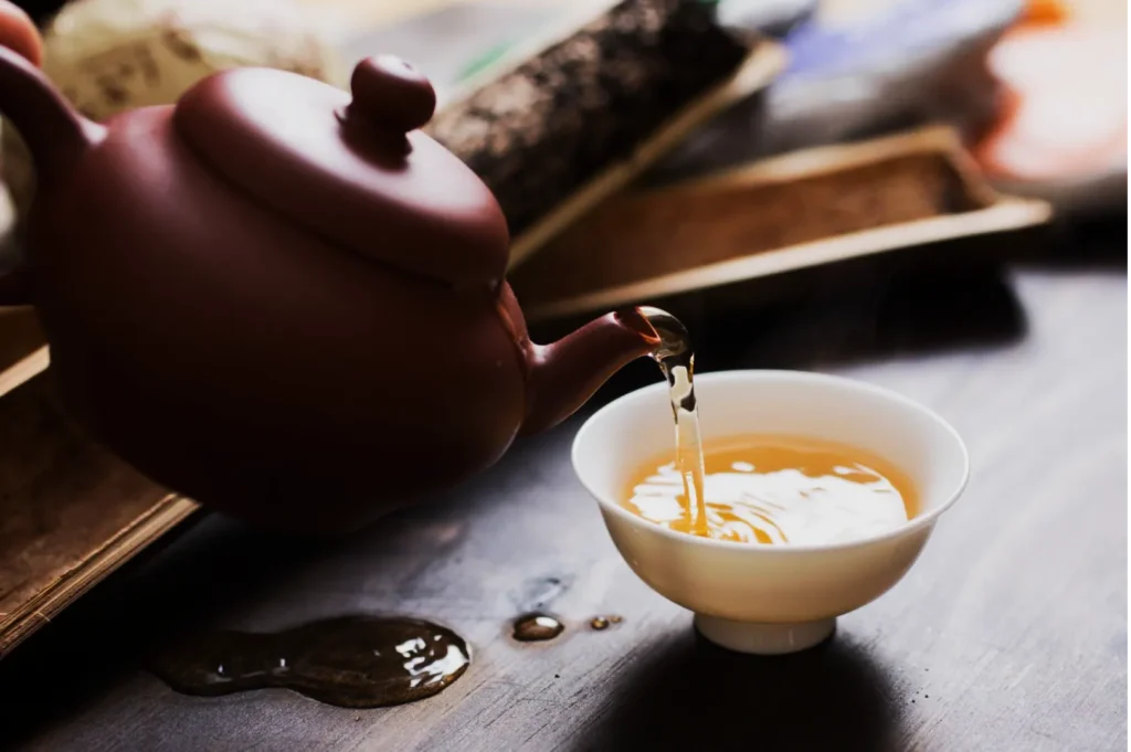 A brown teapot pours steaming tea into a white teacup on a wooden table, with spilled droplets nearby, creating a warm, inviting atmosphere.