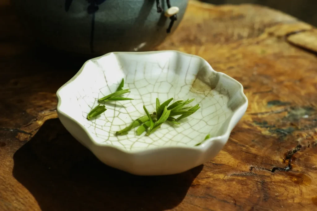 A small, textured white dish with jade-green tea leaves rests on a rustic wooden surface. The scene conveys calm and simplicity.
