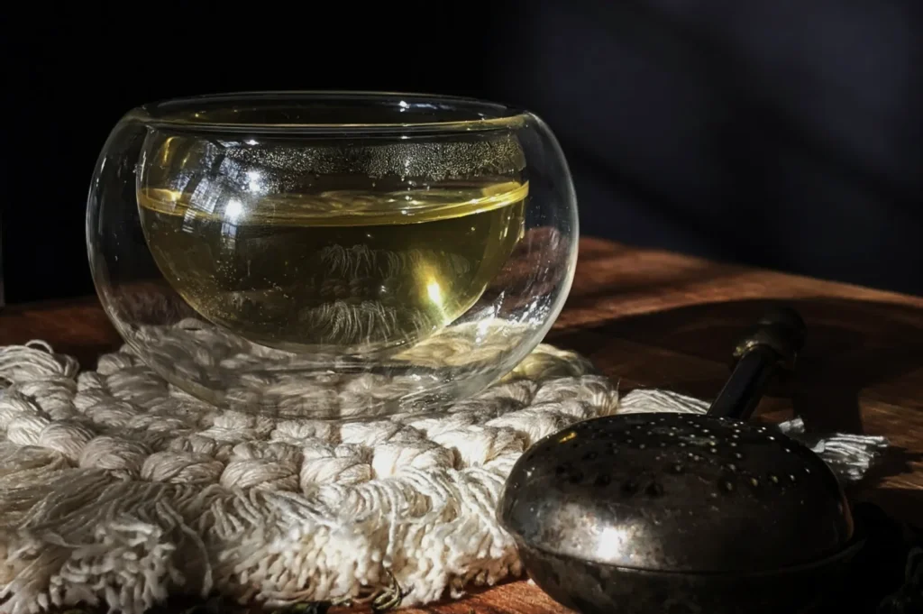 A clear glass cup filled with light green tea sits on a textured, white knitted mat. A vintage, perforated tea infuser rests nearby on the wooden table.