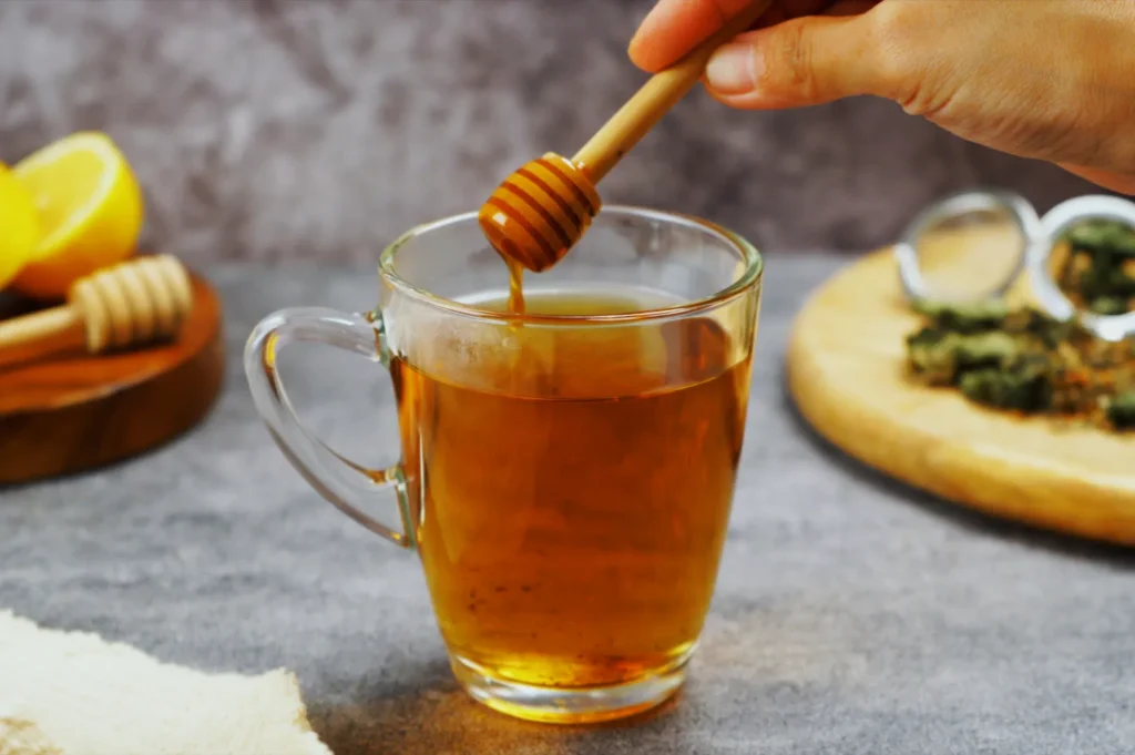 A hand drizzles honey into a clear glass mug of tea. A sliced lemon and herbs are in the background on a wooden board, creating a warm, cozy atmosphere.