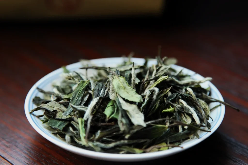 A white ceramic dish filled with dried white tea leaves sits on a wooden surface. The leaves are elongated and slightly curled, with a subtle green and silver hue, evoking a sense of natural and fresh delicacy.