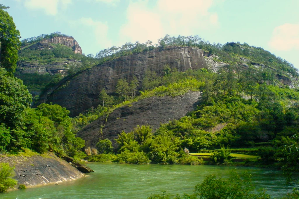 Lush green hills rise steeply beside a calm river under a bright blue sky. Dense vegetation covers rocky slopes, creating a serene, natural landscape.