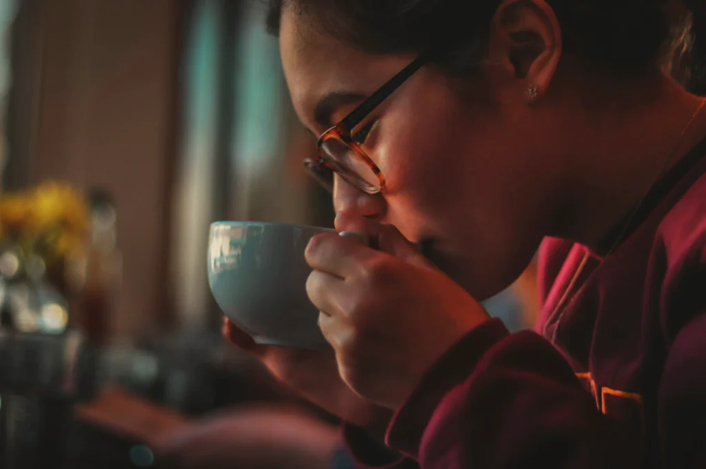 A person in glasses sips from a gray mug, wearing a maroon sweater in a cozy, softly lit café. The mood is warm and relaxed.