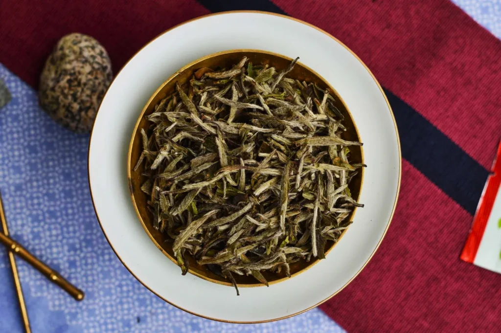 A bowl of dried white tea leaves sits on a white plate, atop a red and blue fabric background. Nearby are a speckled stone and golden chopsticks.