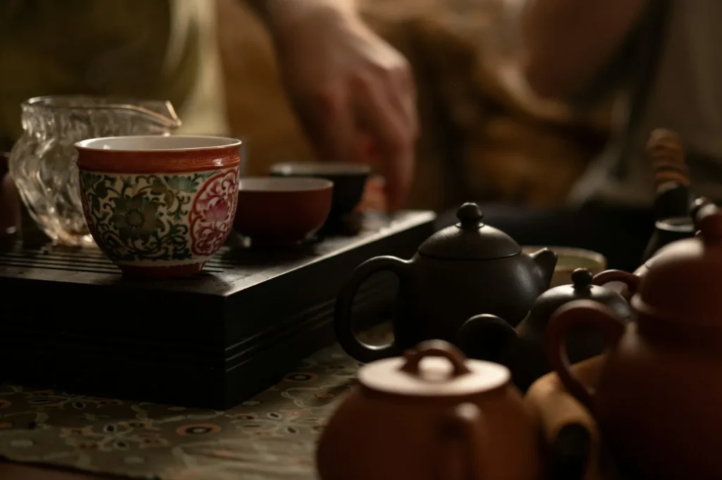 A cozy tea setting with ornate cups and dark teapots on a wooden tray. A hand reaches out, creating an inviting, warm atmosphere.