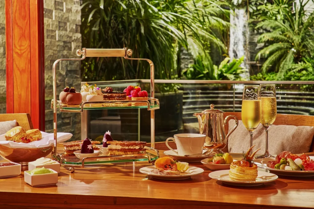 Elegant afternoon tea setting with a two-tiered tray of pastries, scones, and desserts. Champagne flutes, a teapot, and greenery in the background convey a luxurious atmosphere.