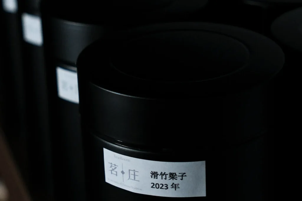 Close-up of a row of black cylindrical canisters with white labels. The label on the front canister reads "2023" in Japanese characters. Dim lighting.