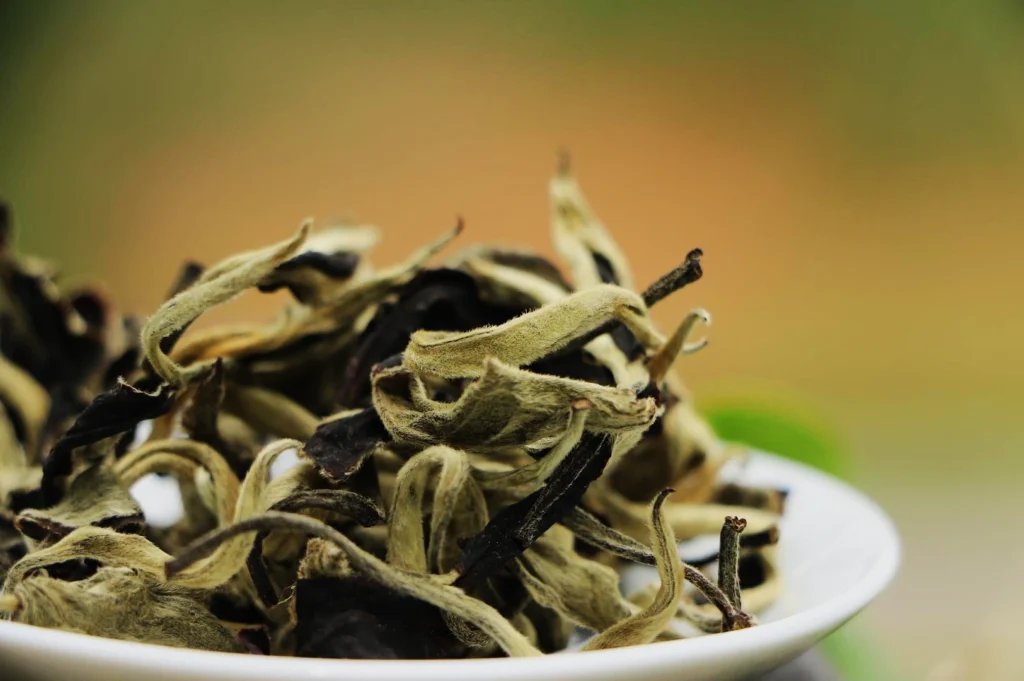 Close-up of dry black and green tea leaves in a white bowl, set against a soft, out-of-focus green and orange background, conveying a natural and earthy feel.