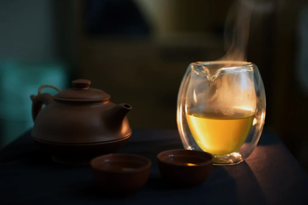 A warm scene showing a steaming glass cup of tea beside a brown teapot and two cups on a dark surface. The ambiance is cozy and inviting.