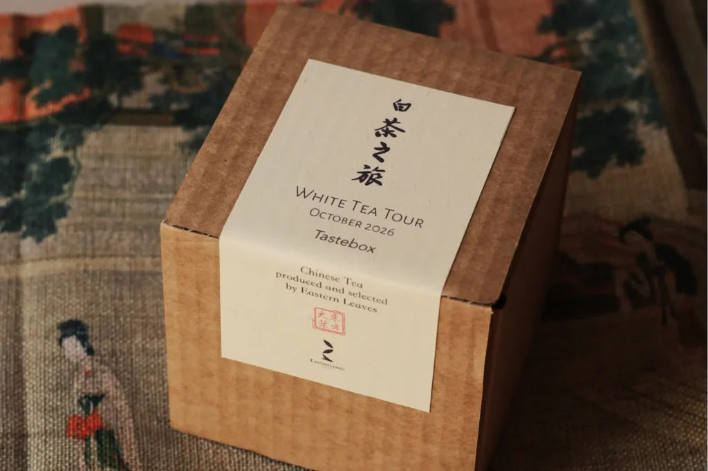 A cardboard box labeled "White Tea Tour, October 2026, Tastebox" sits on a patterned fabric with traditional Asian artwork, evoking a sense of cultural elegance.