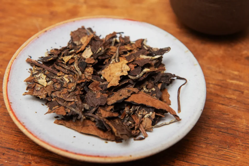 A small, white plate holds a pile of dried, brown tea leaves with visible stems, placed on a wooden table, creating a rustic and earthy ambiance.