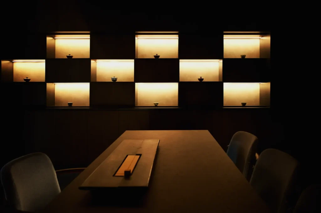 Dimly lit room featuring a modern shelf with illuminated cubbyholes displaying small vases. A sleek wooden table with chairs adds a minimalist touch.