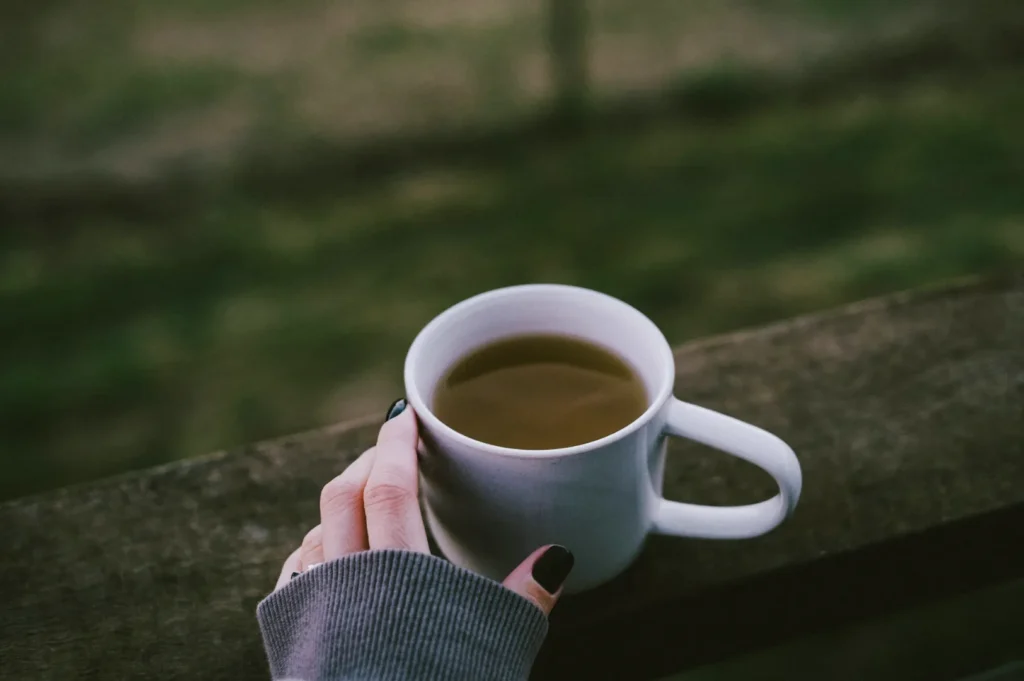 A hand with dark nail polish holds a steaming white mug of tea, resting on a wooden railing. The background shows a blurred, serene green landscape.